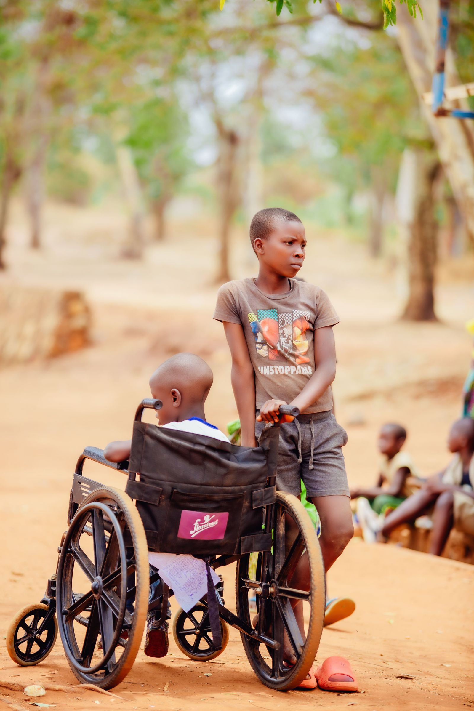 A boy stands beside a younger child seated in a wheelchair on a sandy path.